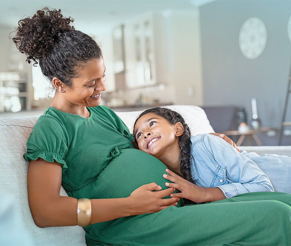 Daughter gently touching her mother’s pregnant belly while sitting together on the couch in their cozy living room